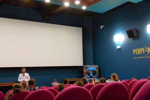 Cinéma intime : sièges roses, écran blanc, murs bleus ; animateur échange avec enfants devant " PERPIGNAN ".