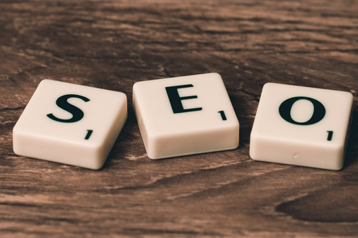 Trois tuiles Scrabble blanches forment "SEO" sur bois, évoquant l'optimisation de la présence digitale.